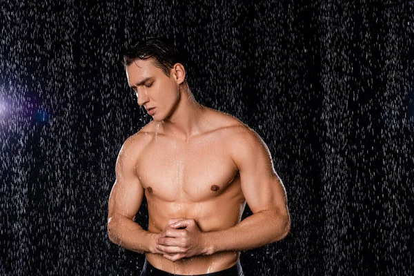 Shirtless man with closed eyes and clenched hands posing under rain on black background — Stock Photo