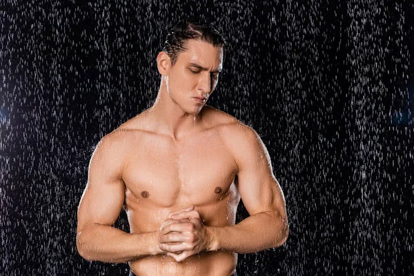 Man with muscular body and clenched hands standing under water drops on black background — Stock Photo