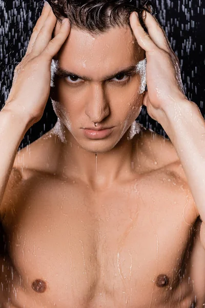 Без сорочки чоловік торкається голови, дивлячись на камеру під душем на чорному тлі — стокове фото