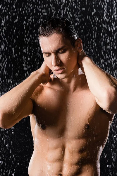 Hombre sin camisa con los ojos cerrados posando bajo la ducha sobre fondo negro - foto de stock