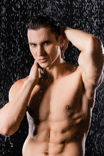 Homem com corpo perfeito tocando pescoço e olhando para a câmera sob o chuveiro no fundo preto — Fotografia de Stock