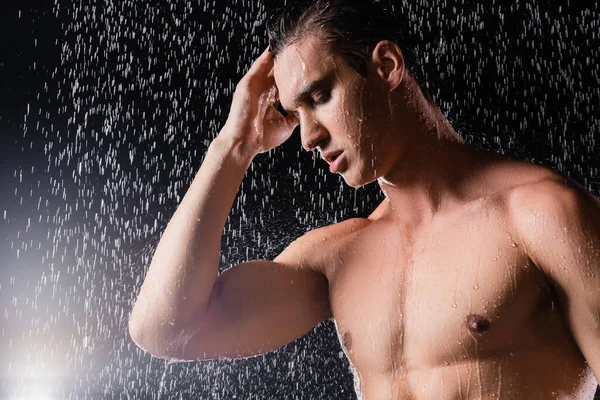 Hombre sin camisa con los ojos cerrados tocando la cabeza mientras se ducha sobre fondo negro - foto de stock