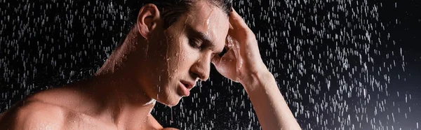 Man with closed eyes touching hair while showering on black background, banner — Stock Photo