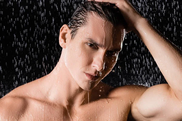 Wet man with bare shoulders looking at camera under shower on black background — Stock Photo