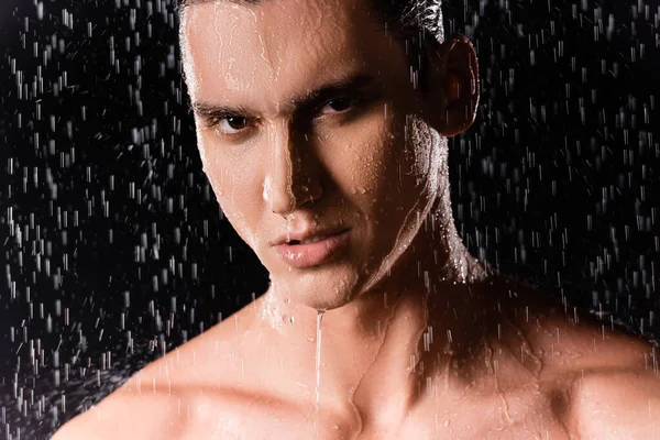 Portrait of man with bare shoulders looking at camera under shower on black background — Stock Photo