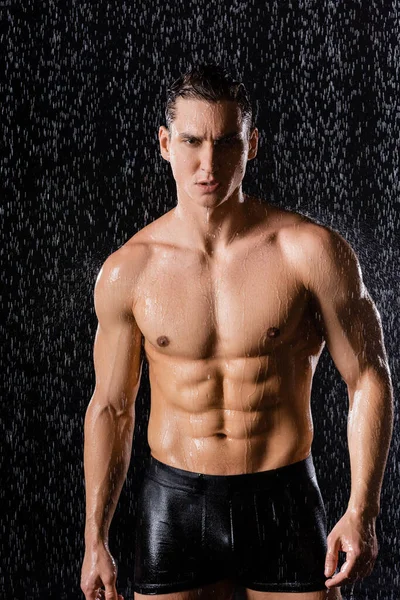 Без рубашки мускулистый мужчина в трусах смотрит в камеру под дождем на черном фоне — стоковое фото