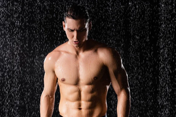 Wet man with perfect body standing under falling water drops on black background — Stock Photo