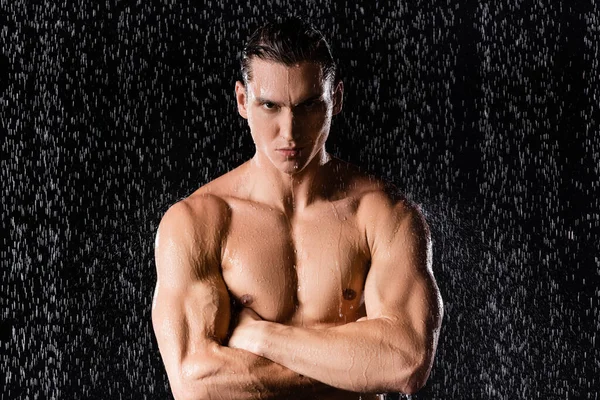 Confident man with muscular torso standing with crossed arms under rain on black background — Stock Photo