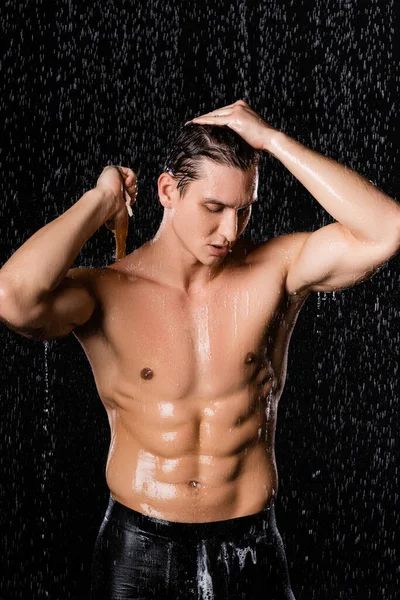 Muskulöser Mann mit geschlossenen Augen, der beim Duschen auf schwarzem Hintergrund die Haare berührt — Stockfoto