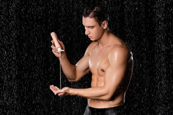 Wet man with muscular torso holding shower gel on black background — Stock Photo