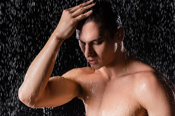 Hombre con los ojos cerrados tocando el pelo mientras se ducha sobre fondo negro - foto de stock