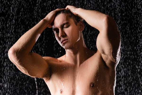 Muscular man with closed eyes and hand on head under refreshing shower on black background — Stock Photo