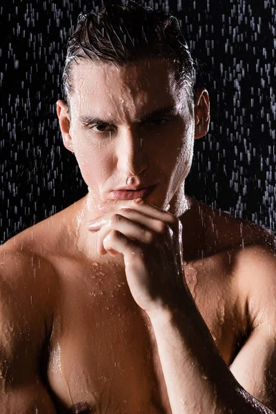 Retrato do homem muscular tocando queixo e olhando para a câmera sob o chuveiro no fundo preto — Fotografia de Stock