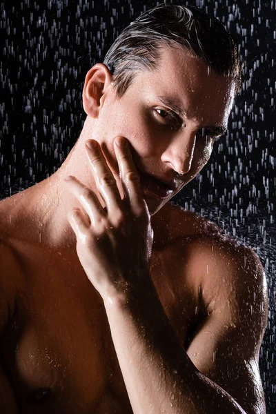 Wet man touching face and looking at camera while showering on black background — Stock Photo