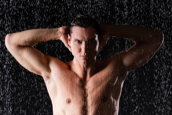 Hombre mojado posando con las manos detrás de la cabeza bajo gotas de agua sobre fondo negro - foto de stock