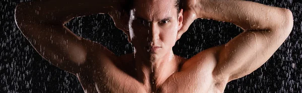 Homem muscular posando com as mãos atrás da cabeça sob o chuveiro no fundo preto, banner — Fotografia de Stock