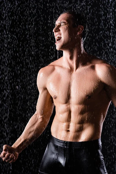 Strong man in underpants singing under rain shower on black background — Stock Photo