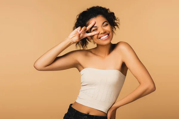 Cheerful african american woman with bare shoulders showing peace sign isolated on beige — Stock Photo