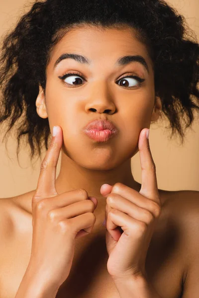 Curly african american woman with bare shoulders touching and pouting cheeks isolated on beige — Stock Photo