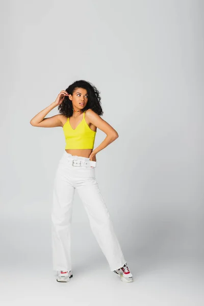 Longitud completa de la mujer afroamericana en traje de moda y elegantes zapatillas de deporte posando con la mano en la cadera aislado en gris - foto de stock