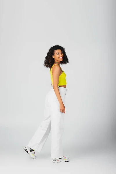 Full length of happy african american woman in trendy outfit and stylish sneakers posing isolated on grey — Stock Photo