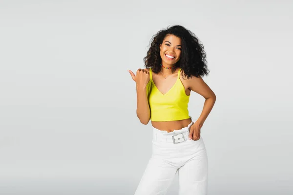 Heureuse femme afro-américaine en haut de culture jaune posant avec la main dans la poche et pointant du doigt avec le pouce isolé sur gris — Photo de stock