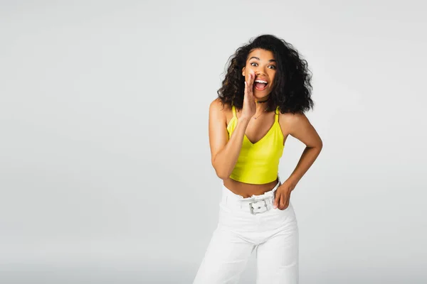 Happy african american woman in yellow crop top posing with hand in pocket and gossiping isolated on grey — Stock Photo