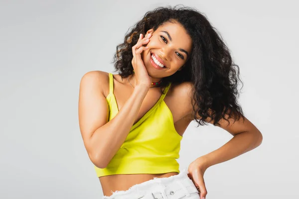 Happy african american woman in yellow crop top posing with hand on hip isolated on grey — Stock Photo