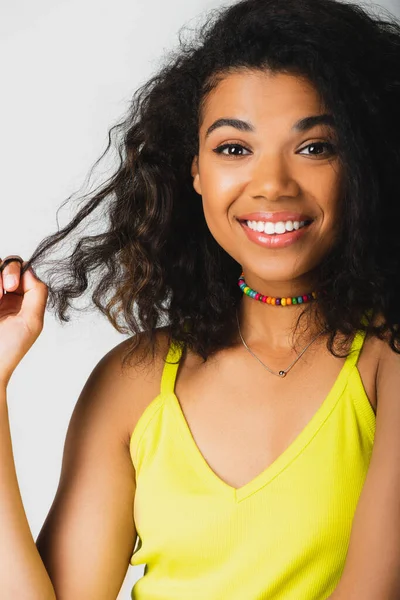 Весела афроамериканка тягне кучеряве волосся і посміхається ізольовано на сірому — стокове фото