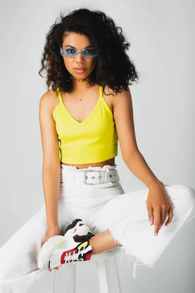 Jolie femme afro-américaine aux lunettes de soleil bleues et tenue tendance posant sur tabouret haut blanc isolé sur gris — Photo de stock