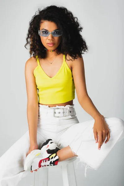 Mujer afroamericana bonita en gafas de sol azules y traje de moda posando en taburete alto blanco aislado en gris - foto de stock