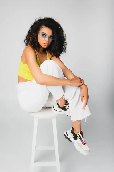 Кудрявая африканская американка в стильных солнцезащитных очках и модном наряде позирует на белом высоком стуле, изолированном на сером — стоковое фото