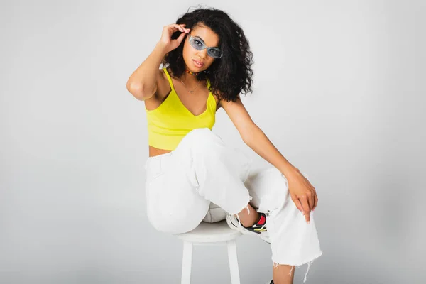 Femme afro-américaine bouclée dans des lunettes de soleil élégantes et tenue tendance posant sur chaise haute blanche sur gris — Photo de stock
