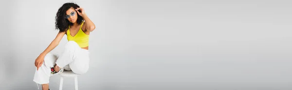 Femme afro-américaine bouclée dans des lunettes de soleil élégantes et tenue tendance posant sur chaise haute blanche sur gris, bannière — Photo de stock