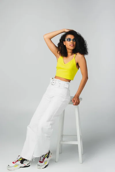 Young african american woman in blue sunglasses and stylish outfit leaning on high stool on grey — Stock Photo