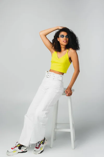 Joven mujer afroamericana en gafas de sol azules y elegante atuendo apoyado en taburete alto en gris - foto de stock