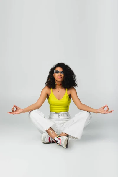 Young african american woman in blue sunglasses and stylish outfit sitting with crossed legs and meditating on grey — Stock Photo