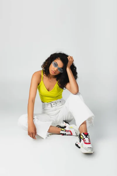 Joven afroamericana americana mujer en azul gafas de sol y elegante traje sentado y posando en gris - foto de stock