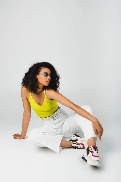 Curly african american woman in blue sunglasses and stylish outfit sitting and posing on grey — Stock Photo
