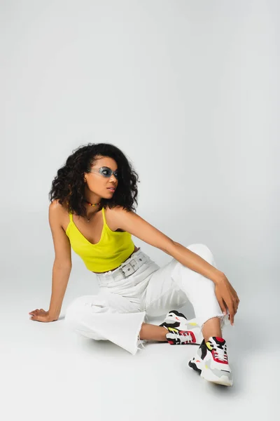 Mujer americana africana rizada en gafas de sol azules y elegante traje sentado y posando en gris - foto de stock