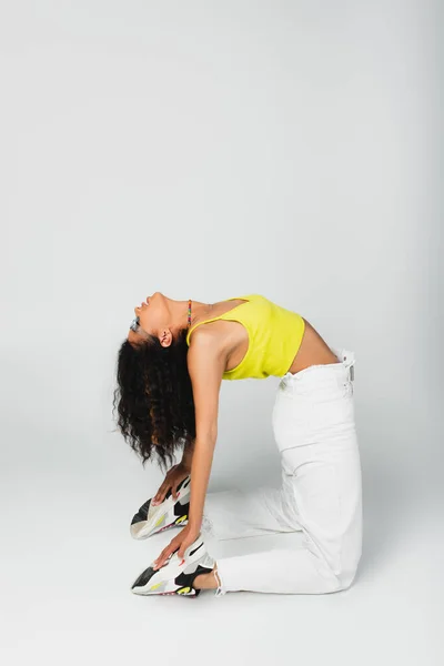 Flexible african american woman in blue sunglasses and stylish outfit posing on grey — Stock Photo