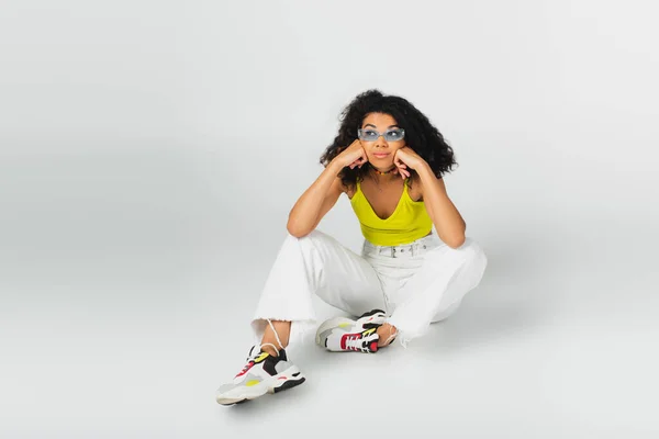Curly african american woman in blue sunglasses and stylish sneakers sitting on grey — Stock Photo