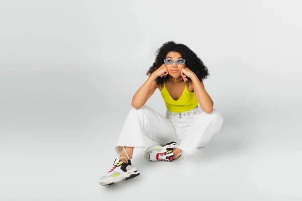 Femme afro-américaine bouclée en lunettes de soleil bleues et baskets élégantes assis sur gris — Photo de stock