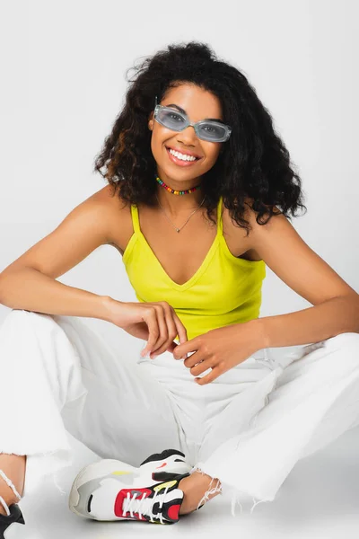 Mulher americana africana alegre em óculos de sol azuis e roupa elegante posando isolado no cinza — Fotografia de Stock