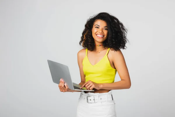 Positive african american freelancer in yellow crop top using laptop isolated on grey — Stock Photo