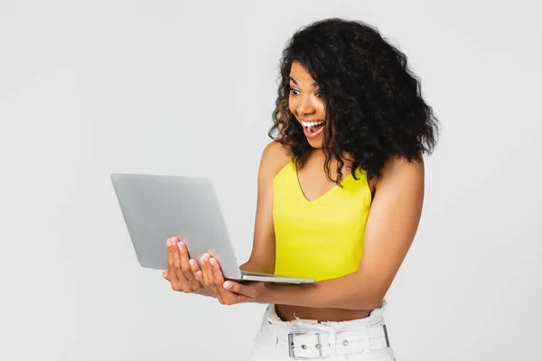 Excited african american freelancer in yellow crop top using laptop isolated on grey — Stock Photo