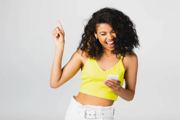 Mujer afroamericana positiva en auriculares inalámbricos sosteniendo el teléfono inteligente y apuntando con el dedo aislado en gris - foto de stock
