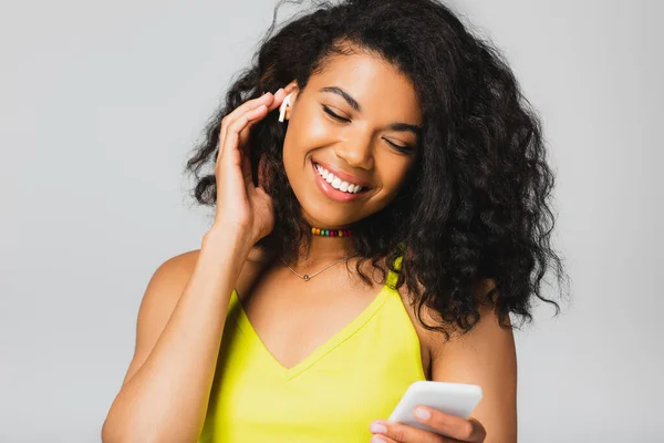 Mujer afroamericana feliz en auriculares inalámbricos sosteniendo teléfono inteligente aislado en gris - foto de stock