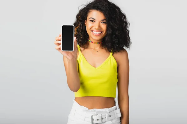 Alegre mujer afroamericana sosteniendo teléfono inteligente con pantalla en blanco aislado en gris - foto de stock