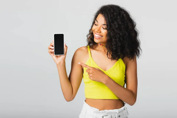 Щаслива афроамериканка вказує на смартфон з порожнім екраном ізольовано на сірому — стокове фото